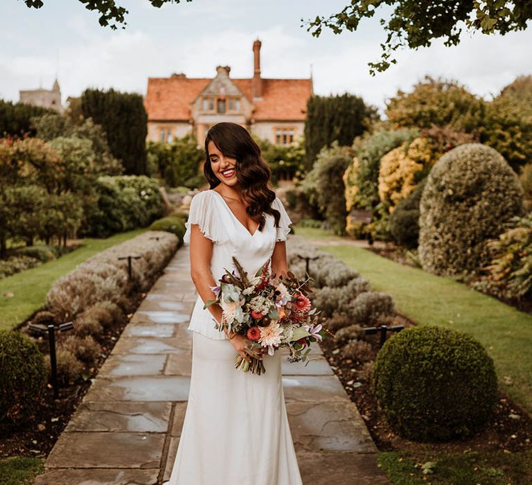 Bride in v-neck satin wedding dress with sheer butterfly sleeves and ruffled layer on top holding flower bouquet with roses, carnations, eucalyptus, baby's-breath and dried flowers 