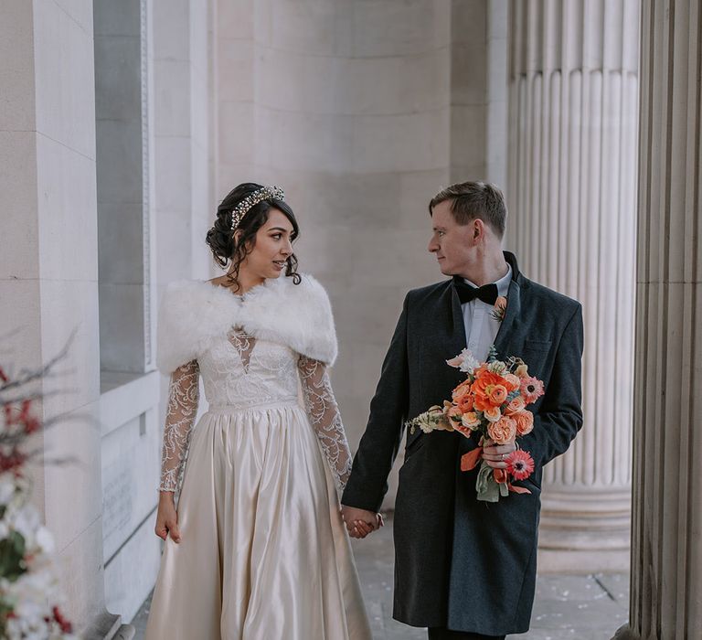 Groom in black overcoat, classic black tuxedo and a black bowtie holding bouquet with light pink poppies, orange garden roses, eucalyptus, Christmas red berry garlands, dried flowers and sage green and orange ribbon with bride in Bride in taffeta material princess wedding dress with long lace sleeves  and cropped faux fur bridal cover up 