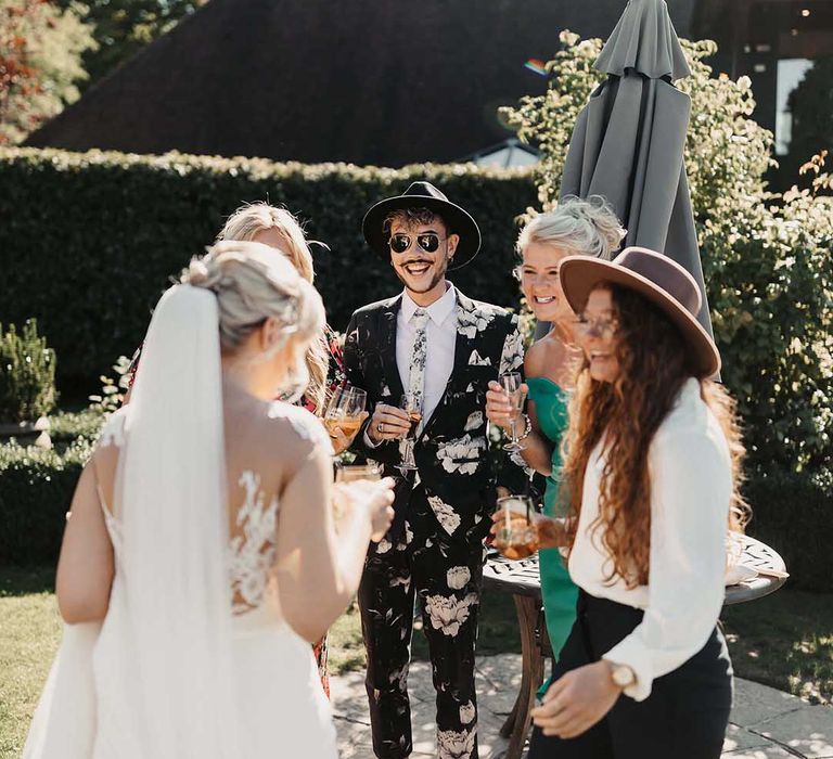 Wedding guest in black and off-white rose patterned suit and matching tie with sunglasses and fedora hat standing with bride in lace wedding dress and other guests drinking champagne 