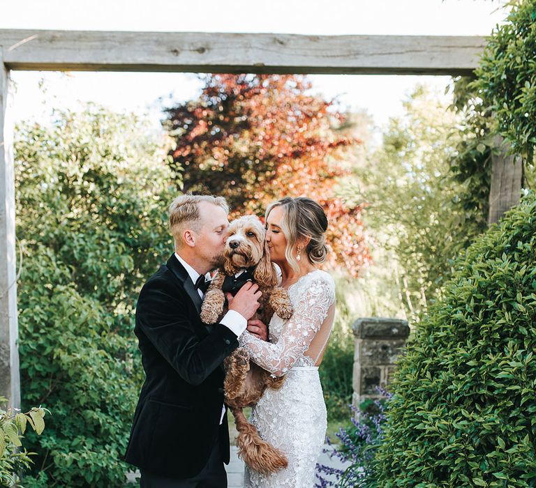 Bride in long sleeve shoulder pad wedding dress and groom in black tuxedo kissing their pet dog 