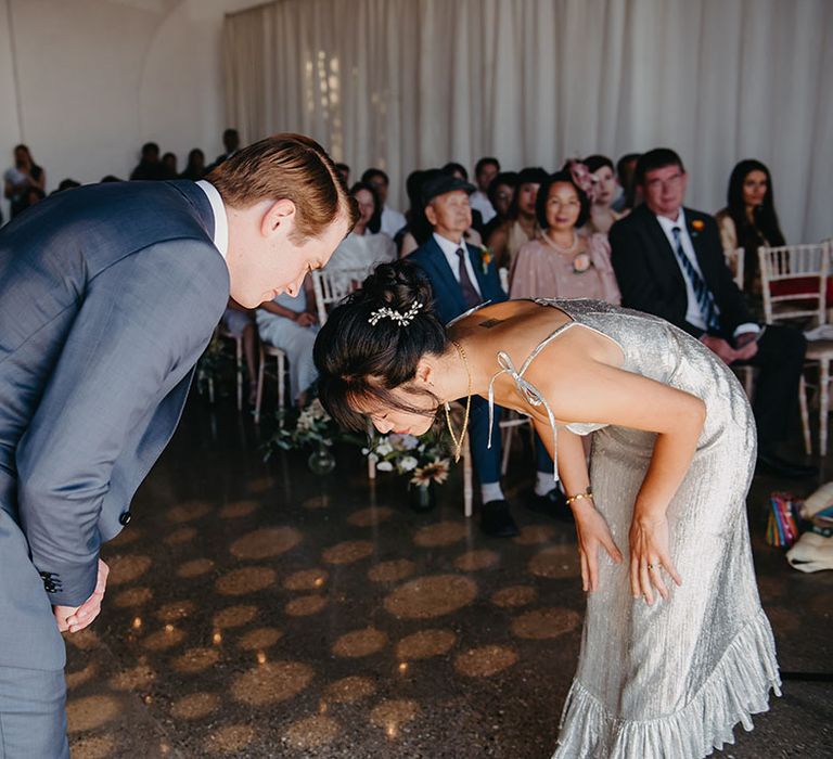 Bride in silver wedding dress & groom bow toward one another during bespoke wedding ceremony 