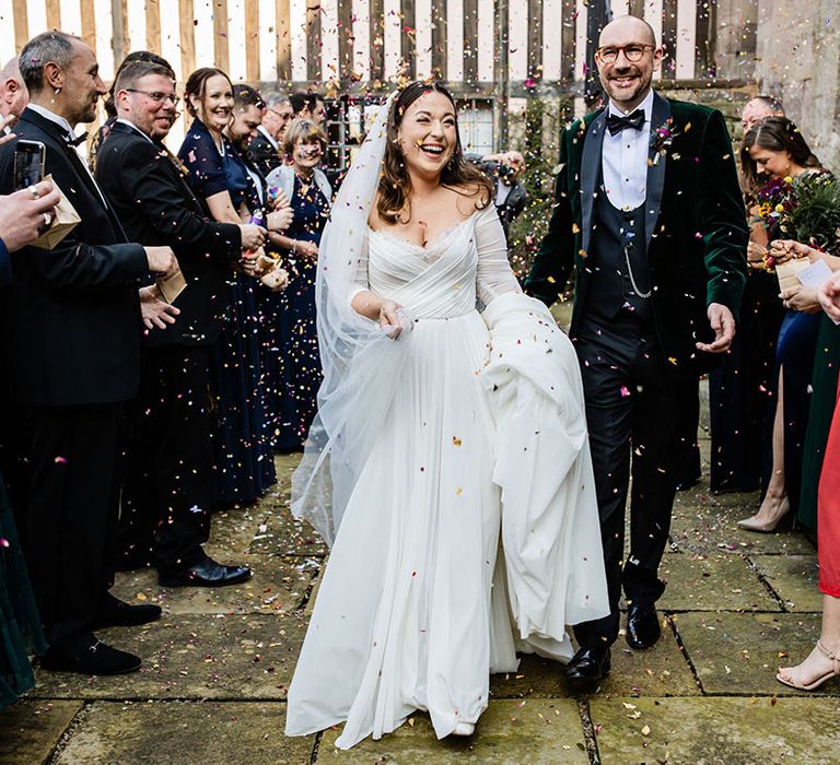 Groom in velvet tuxedo walking with the bride in a tulle wedding dress smiling brightly as they have their confetti moment after their civil ceremony 