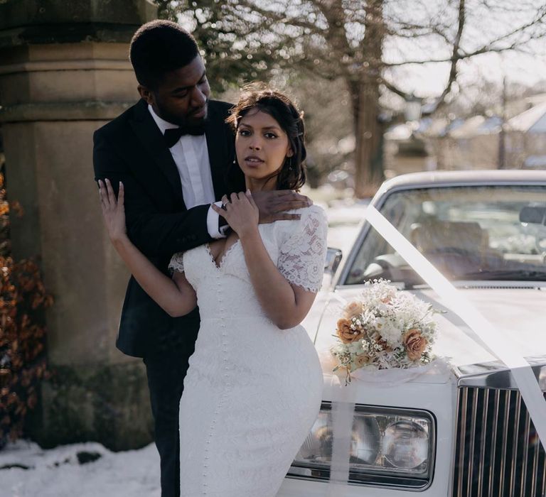 Bride in v neck short sleeve wedding dress with front slit and corset button detailing being embraced by groom in classic black tuxedo, black bowtie and white pocket square 