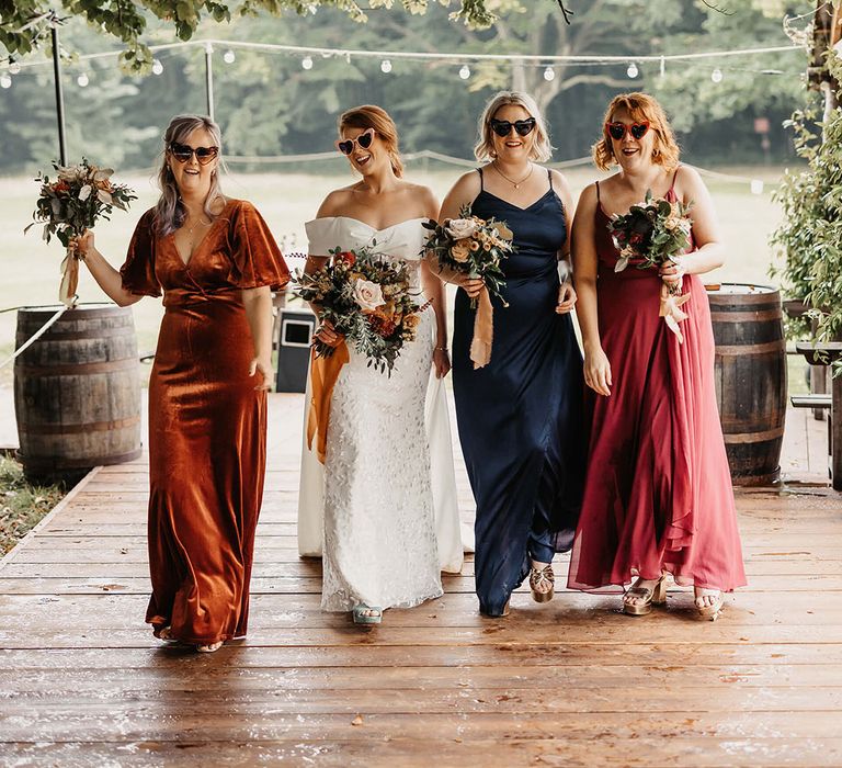 Bride in sunglasses walks alongside her bridesmaids in mismatched bridesmaid dresses in Autumnal colours