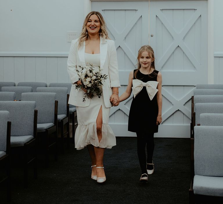 The bride and groom's daughter in black dress with big bow detail walks the bride down the aisle in a white blazer and ruffle wedding dress 