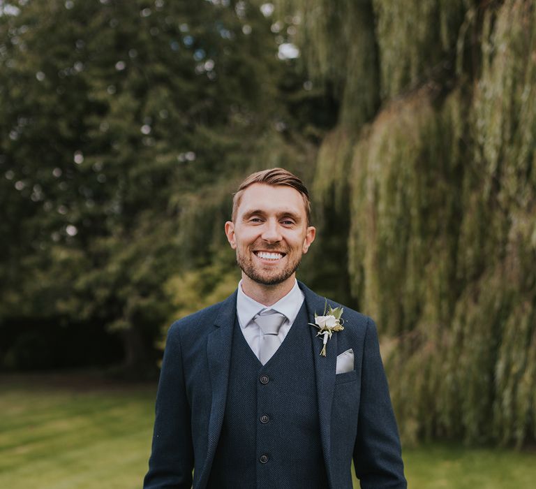 The groom in a three piece dark navy suit with silver grey tie and white rose and gypsophila buttonhole 