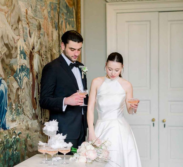 Bride in satin halter neck wedding dress standing with groom in classic black tuxedo standing by champagne tower with candy floss topper 