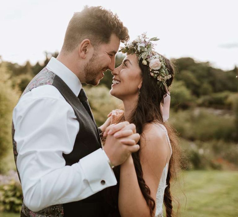 Bride in 3d applique floral sheer overlay wedding dress with white garden rose flower crown and cathedral-length veil embracing groom in dark grey blazer with tartan tie and white garden rose boutonniere 