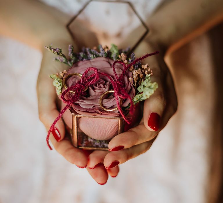 Glass and gold ring box with a dusty pink rose inside and two gold rings tied in red string