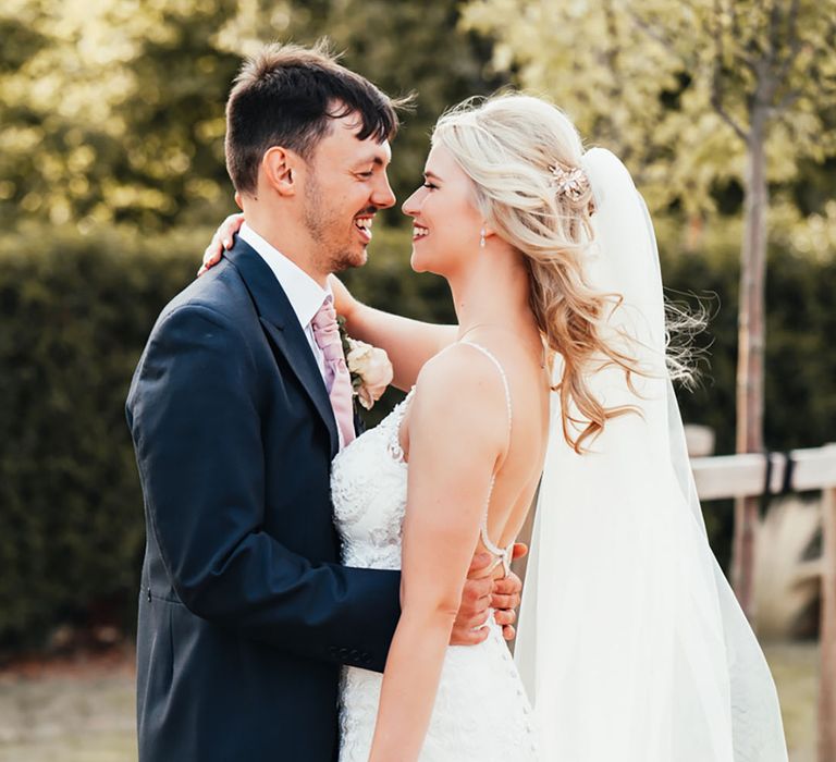 Groom in navy suit with pink tie smiling with the bride in a fitted lace wedding dress with a long veil holding a white wedding bouquet 