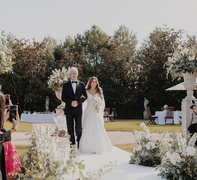 Bride in lace long sleeve Pronovias wedding dress walking down the aisle with carnation, eucalyptus and dried flower aisle flower decorations with father of the bride 