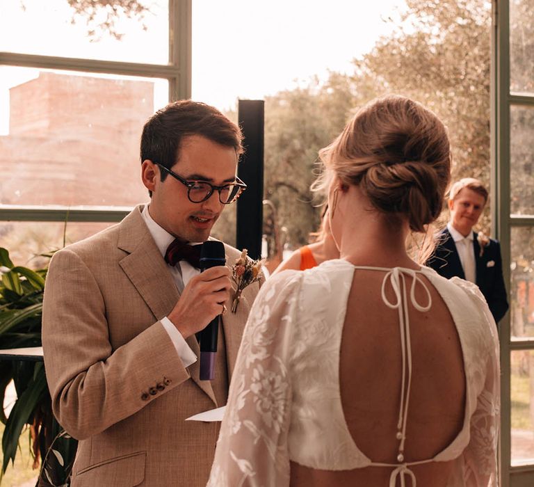 Groom in light cream suit with dried flower buttonhole with tortoise shell glasses reading out his wedding vows to the bride in a Grace Loves Lace bridal two piece for destination wedding 