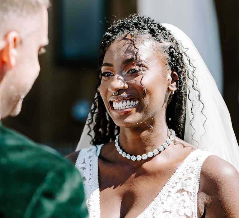 Bride in v neck sleeveless lace Pronovias wedding dress and silver sparkly bridal jewellery standing with groom in bottle green velvet grooms blazer 