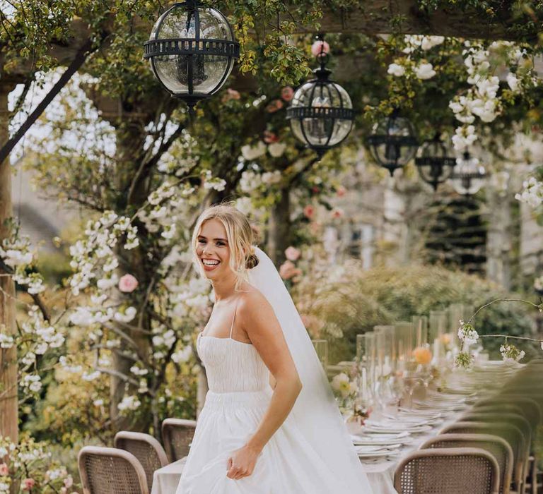 Bride in Watters strappy sleeveless princess wedding dress and chapel length wedding veil standing by outdoor wedding tablescape at Euridge Manor wedding 