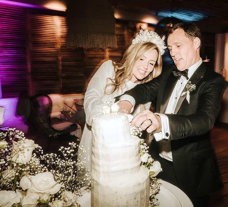 bride in a short sparkly wedding reception dress wearing a dried flower headband cutting the wedding cake with her groom in a tuxedo and bow tie at pure house ibiza venue