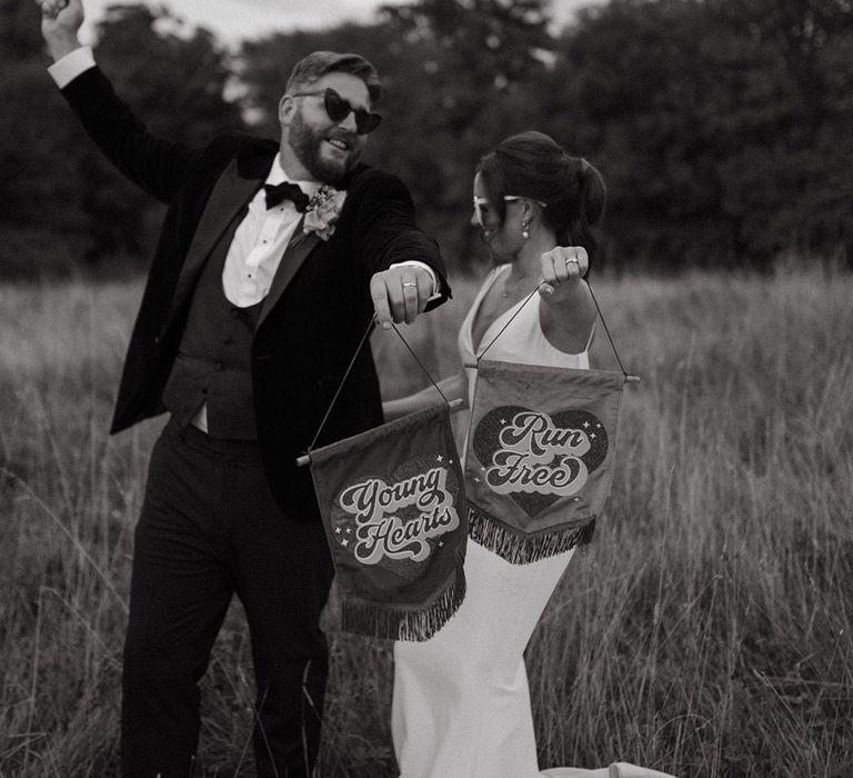 Groom in three piece black tuxedo with the bride in a satin slip dress holding pink and red retro banner signs 