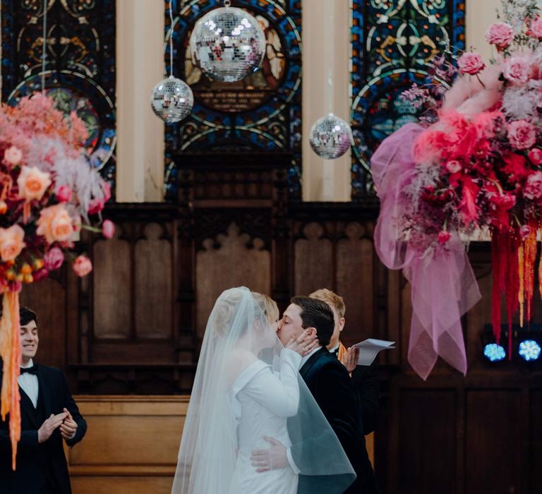 Bride in asymmetrical off shoulder long sleeve satin wedding dress with cathedral length personalised veil kissing groom in classic black suit at the alter of Fabrica Brighton wedding venue