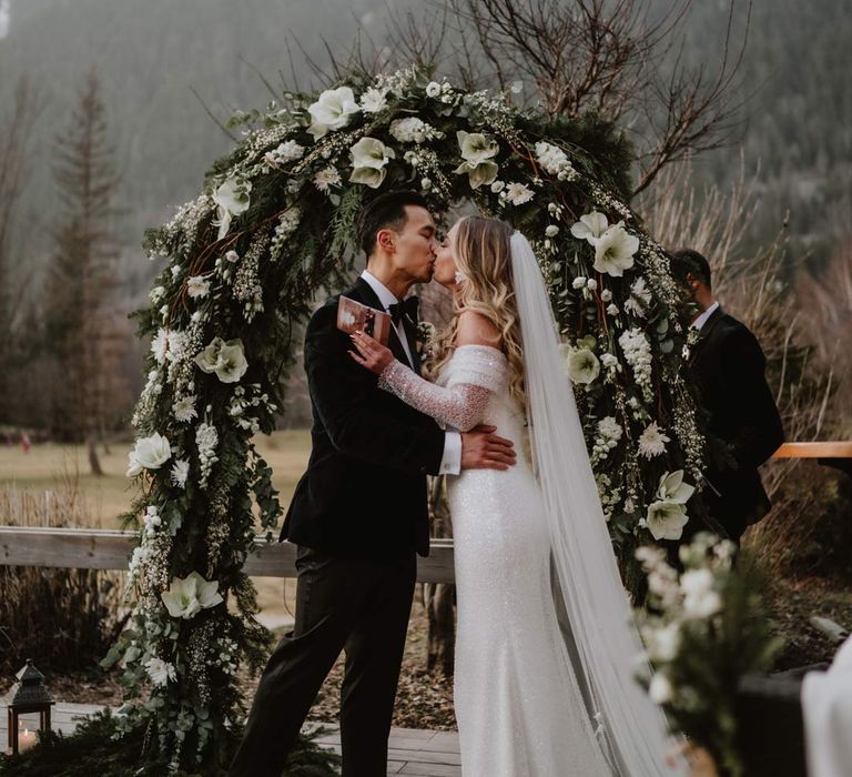 Bride in off shoulder sparkly mermaid wedding dress kissing groom in classic black tuxedo, black bowtie and white garden rose boutonniere standing in front of white garden rose, eucalyptus, baby's-breath and foliage wedding flower arch at French destination ski wedding