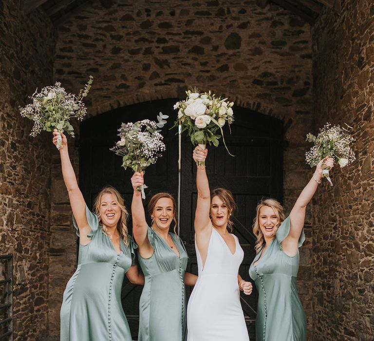 Bridal party in matching satin green dresses with the bride raising their matching white rose and gypsophila bouquets in the air 