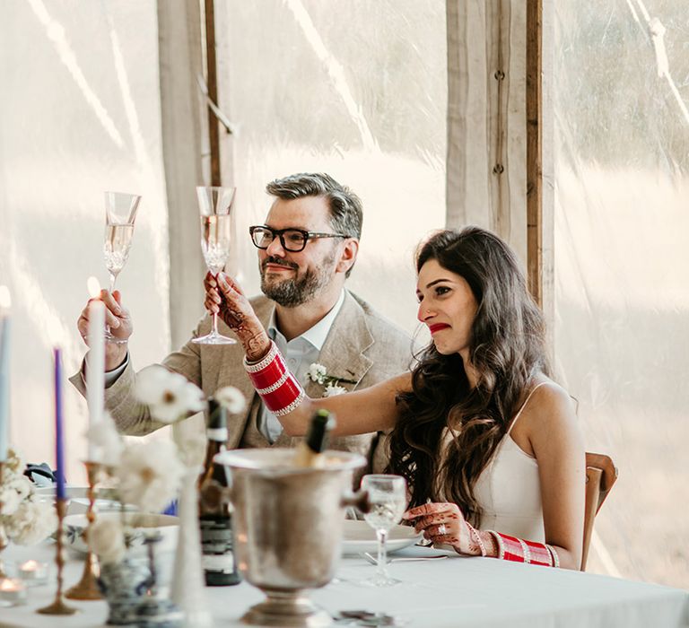 The bride and groom sitting together raise their wine glasses in a toast to one of the wedding speeches 