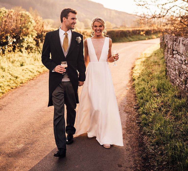 Groom in morning suit with yellow tie holding glass of Guinness walking with bride in Andrea Hawke wedding dress 