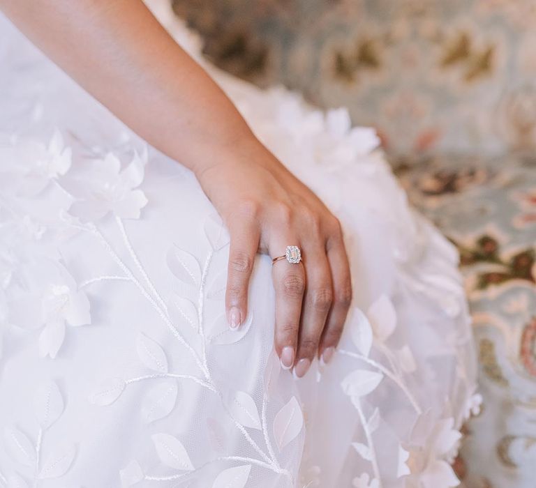 Bride with classic French tip wedding nails with diamond engagement ring resting on floral lace gown 