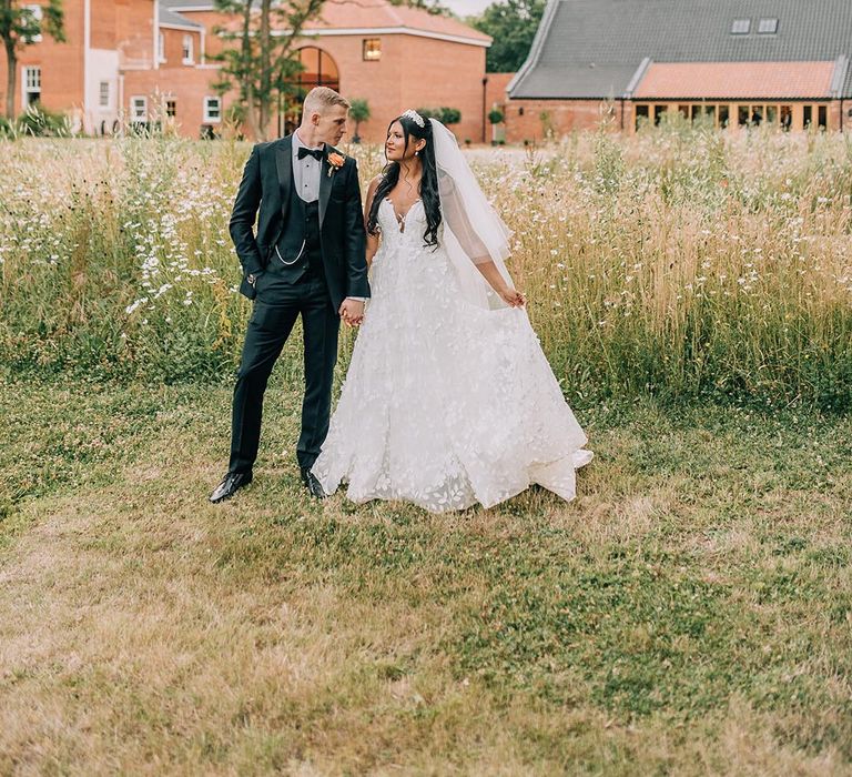 Groom in black tuxedo walking around with the bride in floral lace bridal gown 