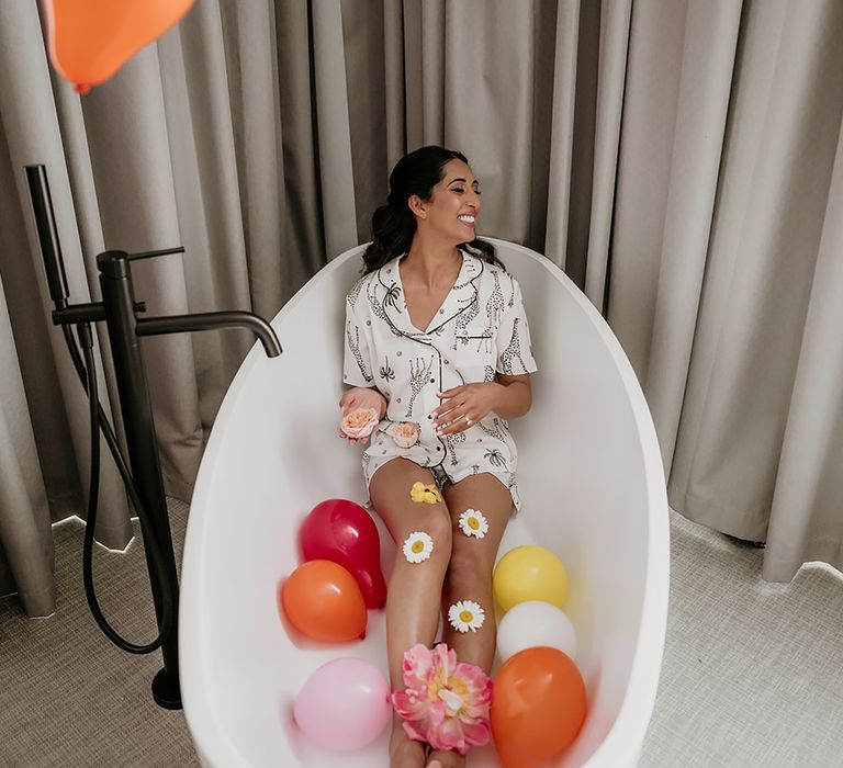 Bride in white giraffe wedding pyjamas sits in bath tub with flowers and balloons