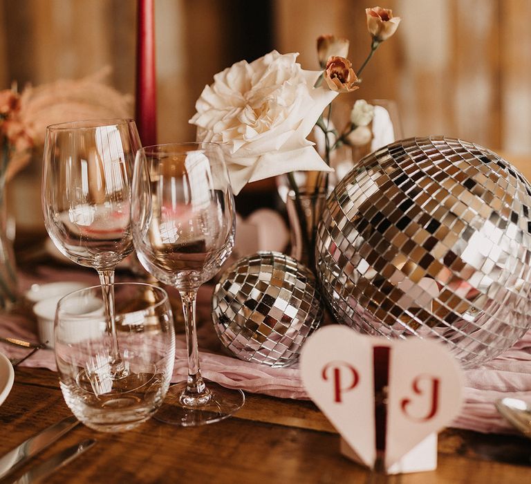 Disco balls with pink table runner with white rose in bud vase with heart shaped stationery and heart shaped sunglasses favours 