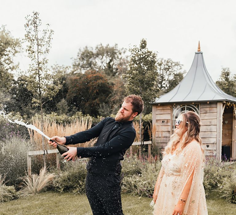 Groom in all black wedding suit pops bottle of champagne with the bride in celebration on their wedding day 