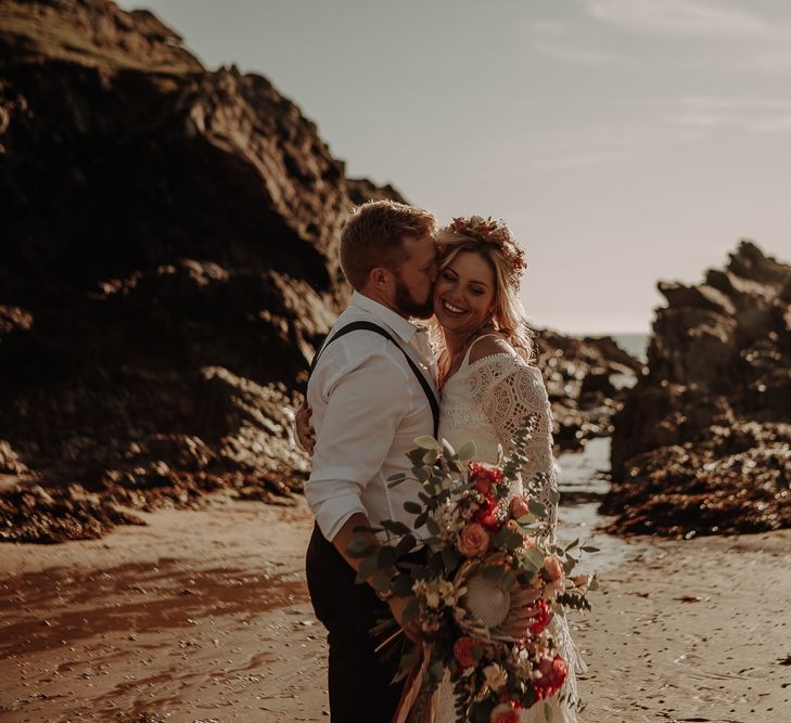 Bride and groom share a kiss on the beach with large autumnal wedding bouquet 