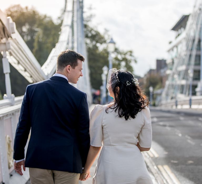 Bride with shoulder length brown hair and birdcage veil holding her grooms hand on a bridge 