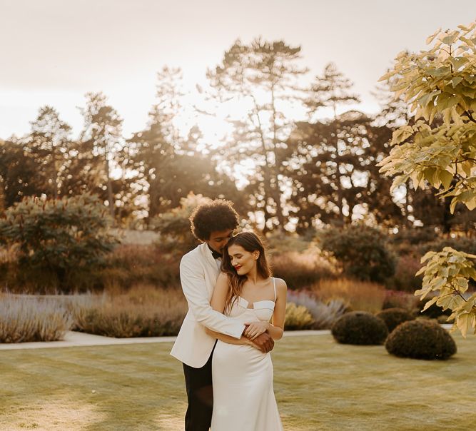 Black groom with afro hair and white tuxedo jacket embracing his bride with long brown hair and red lipstick in a satin wedding dress with cup detail and thin straps 