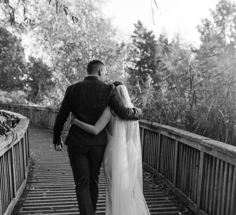 Black and white wedding day photography of bride and groom walking across a bride in the autumnal leaves 