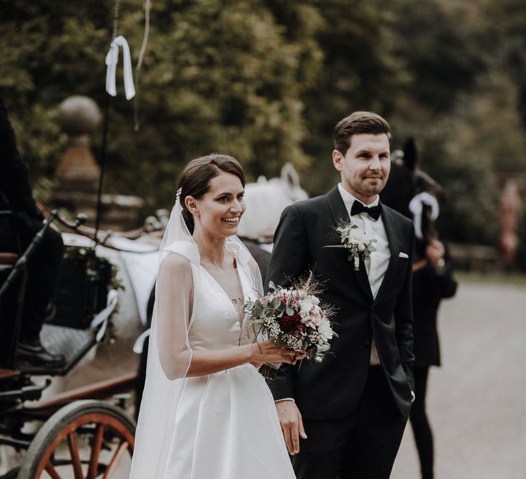 Bride in white Jesus Piero gown and cathedral length veil arrives at German wedding reception with groom in black Tiger of Sweden tux