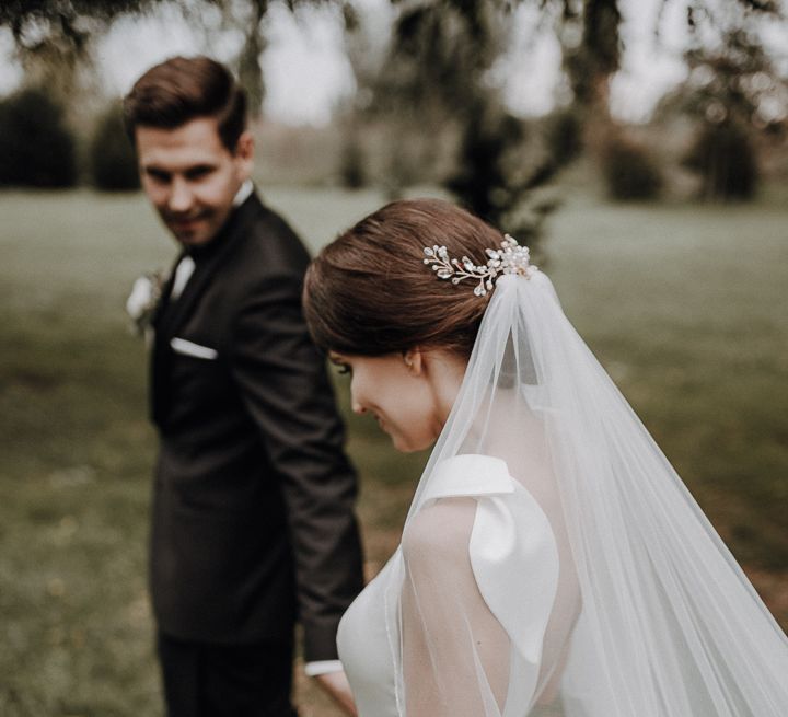 Bride in white Jesus Piero gown and cathedral length veil walks with groom in black Tiger of Sweden tux in gardens at German wedding