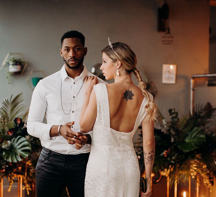 Bride in a jacquard midi dress with low back, sleek ponytail and crown standing next to her groom in black leather trousers and a white shirt 