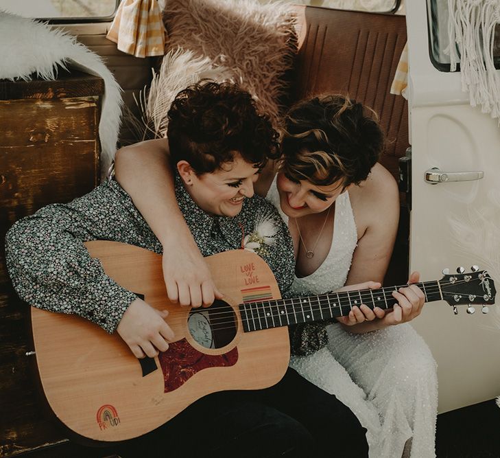 A lesbian couple sit at the door opening of a camper van. One plays the guitar and the other hugs her and laughs.