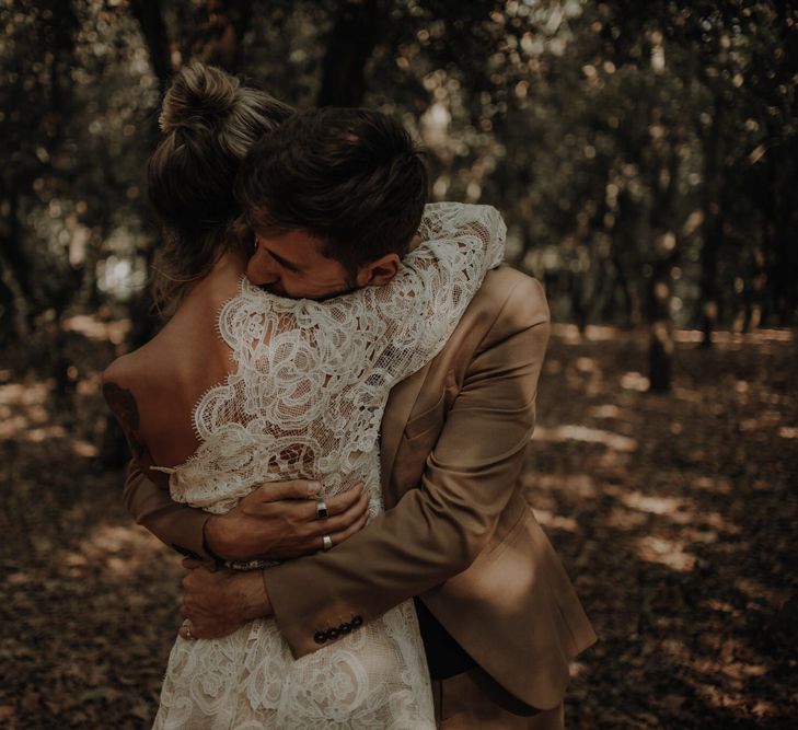 Groom in a beige suit embracing his bride in a one shoulder wedding dress with long lace sleeve