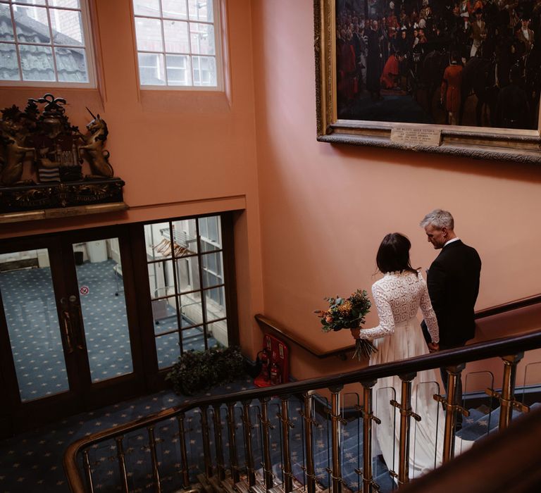 Bride in long sleeved lace top Self Portrait wedding dress holding colourful bouquet walks down Bristol Registry Office stairs holding hands with groom in brown woollen blazer and waistcoat