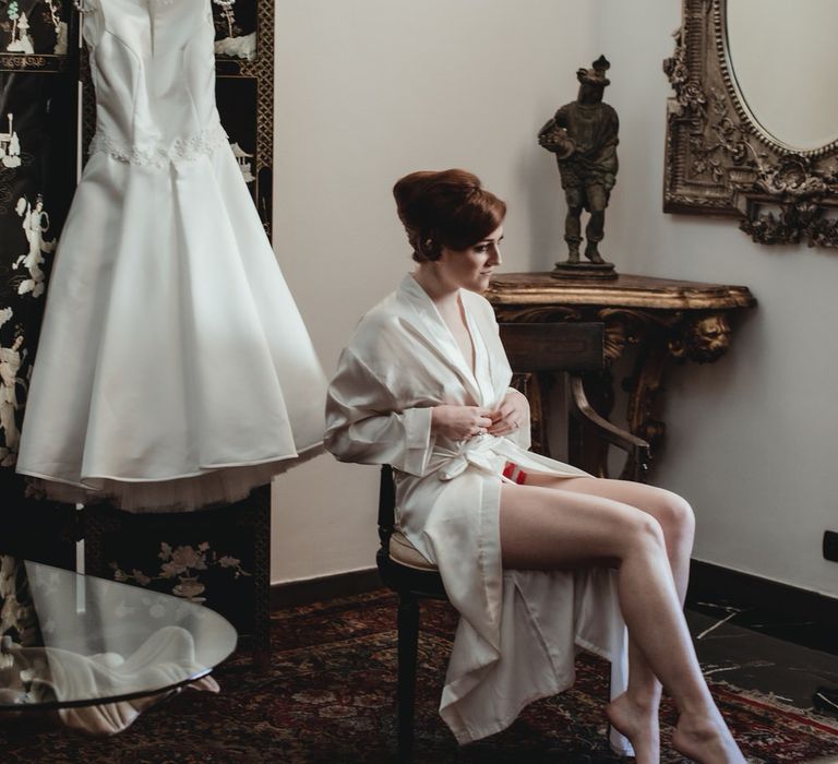 Bride getting ready in a white silk dressing gown, with her vintage style wedding dress hanging behind her