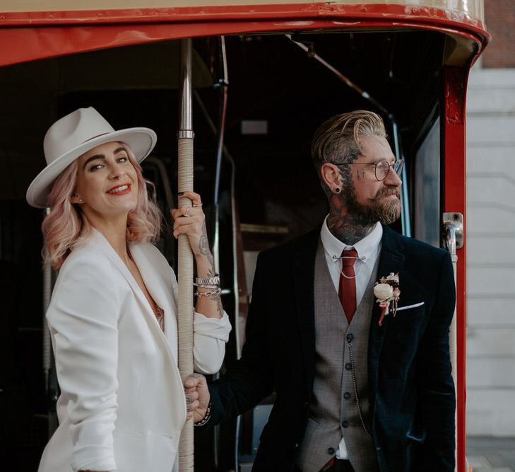 Bride in white suit and fedora holds on to the pole of a red double decker bus whilst standing next to tattooed groom who looks away