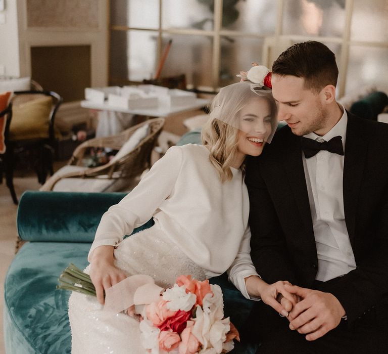 Bride and groom holding hands on a blue velvet sofa, bride is holding a bouquet of pink and white flowers