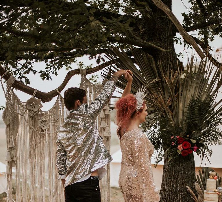 Bride in gold long sleeved sequin wedding dress and white and silver crown headband is spun around by groom in silver sequin suit jacket and black trousers by macrame hanging outside at festival themed wedding