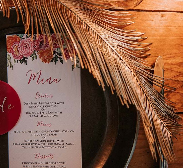 White wedding menu with red calligraphy and tropical floral illustrations on black plate with gold palm leaf decor and red circular table names at festival themed wedding