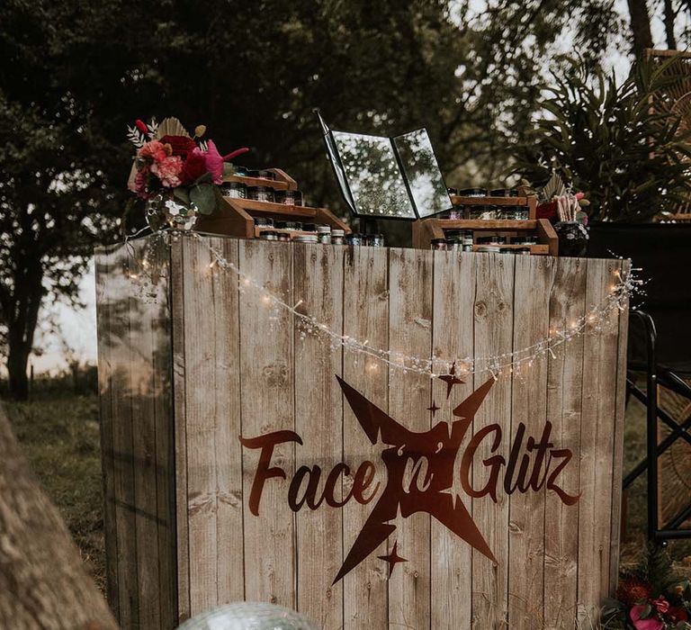 Wooden Face n Glitz glitter station outside at festival themed wedding with disco balls, tropical flower decor and fairy lights 