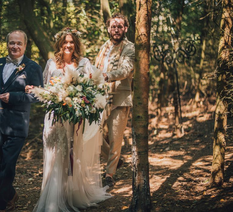 Bride walks through trees with father whilst holding large floral bouquet