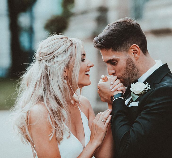 Groom in a black tuxedo with white rose buttonhole flower kissing his brides hand in a slip wedding dress at their Gas Street Church wedding 