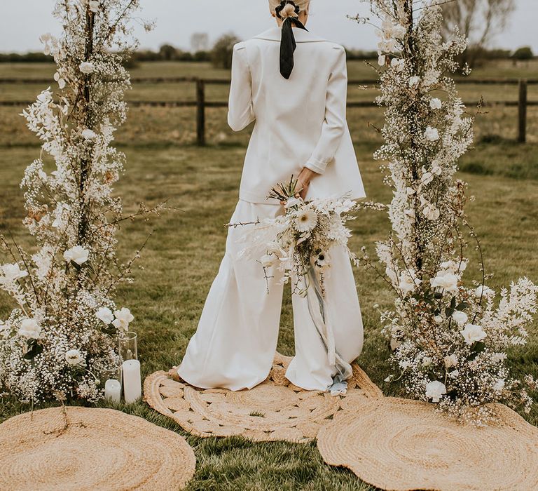 Bride in a white suit with a bun tied in black ribbon standing on a wicker rug under a white flower arch 