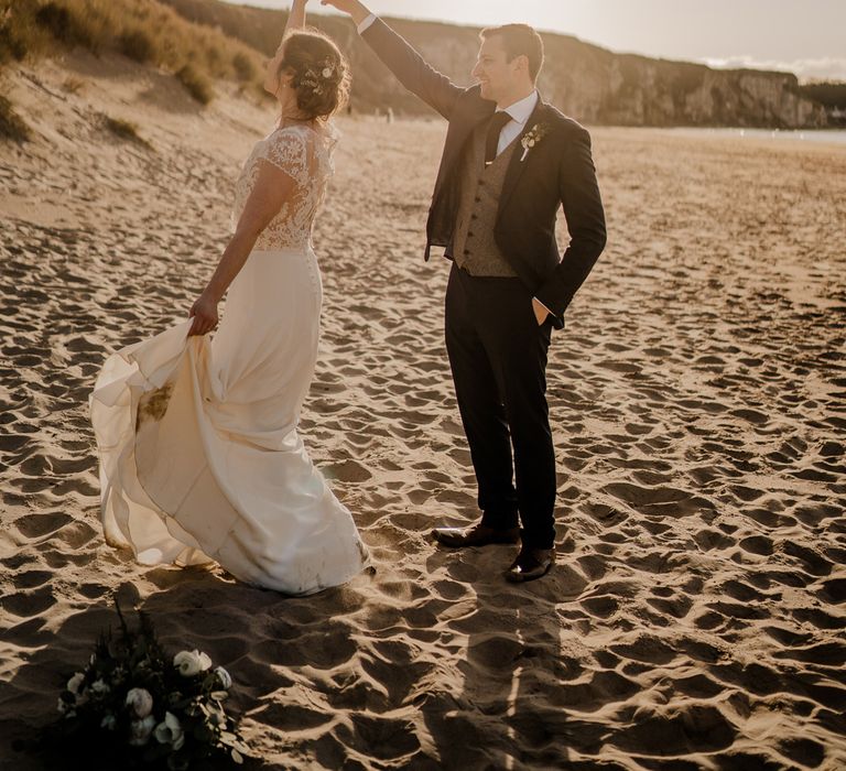 Groom in navy suit twirls around bride in lace top capped sleeve wedding dress with satin skirt on the beach after Dunluce Castle wedding